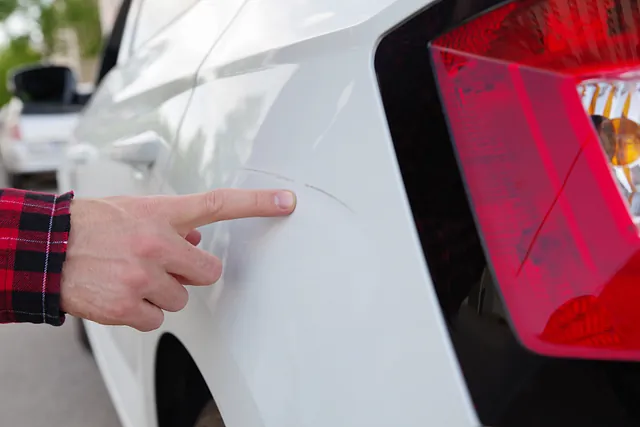 Eine Hand zeigt einen Kratzer auf einem weißen Auto. Der Kratzer kann mithilfe der Smart Repair in Bergisch Gladbach entfernt werden.